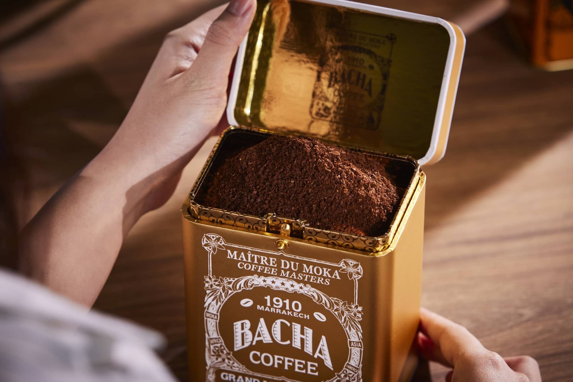 A gold Bacha Coffee tin opened on ground coffee, held in two hands above a counter