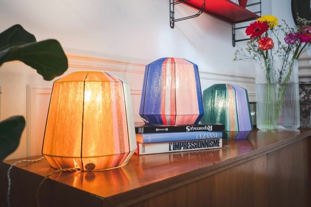 Trois lampes en fil coloré posées sur une enfilade en bois avec livres et fleurs