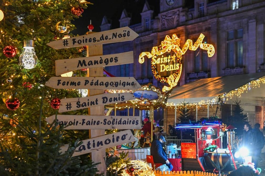 Panneau de bois de Bel Hiver indiquant foires de Noël, manèges, ferme pédagogique et patinoire, sous les guirlandes