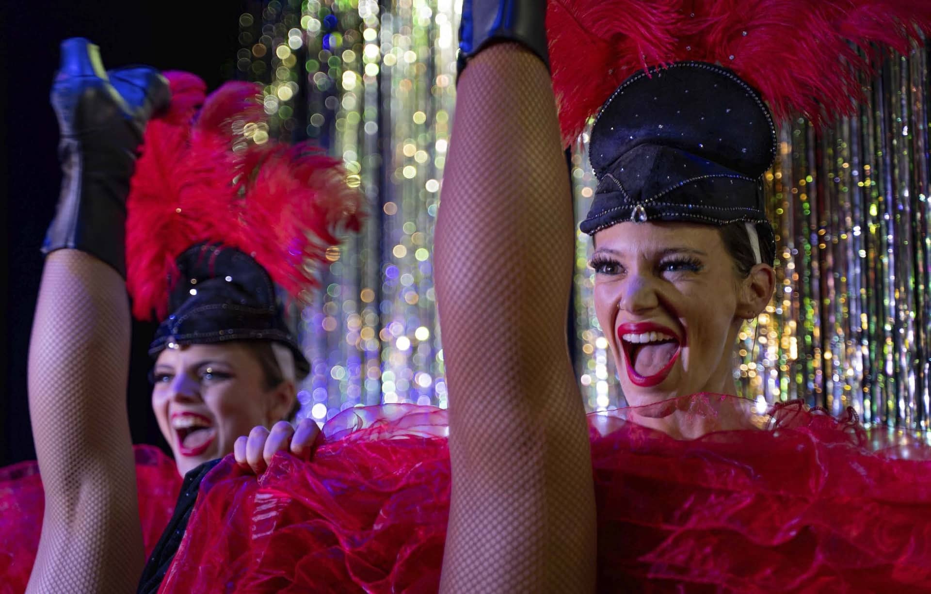 Deux danseuses de revue en plumes rouges et casquettes à paillettes, jambe levée, en plein rire