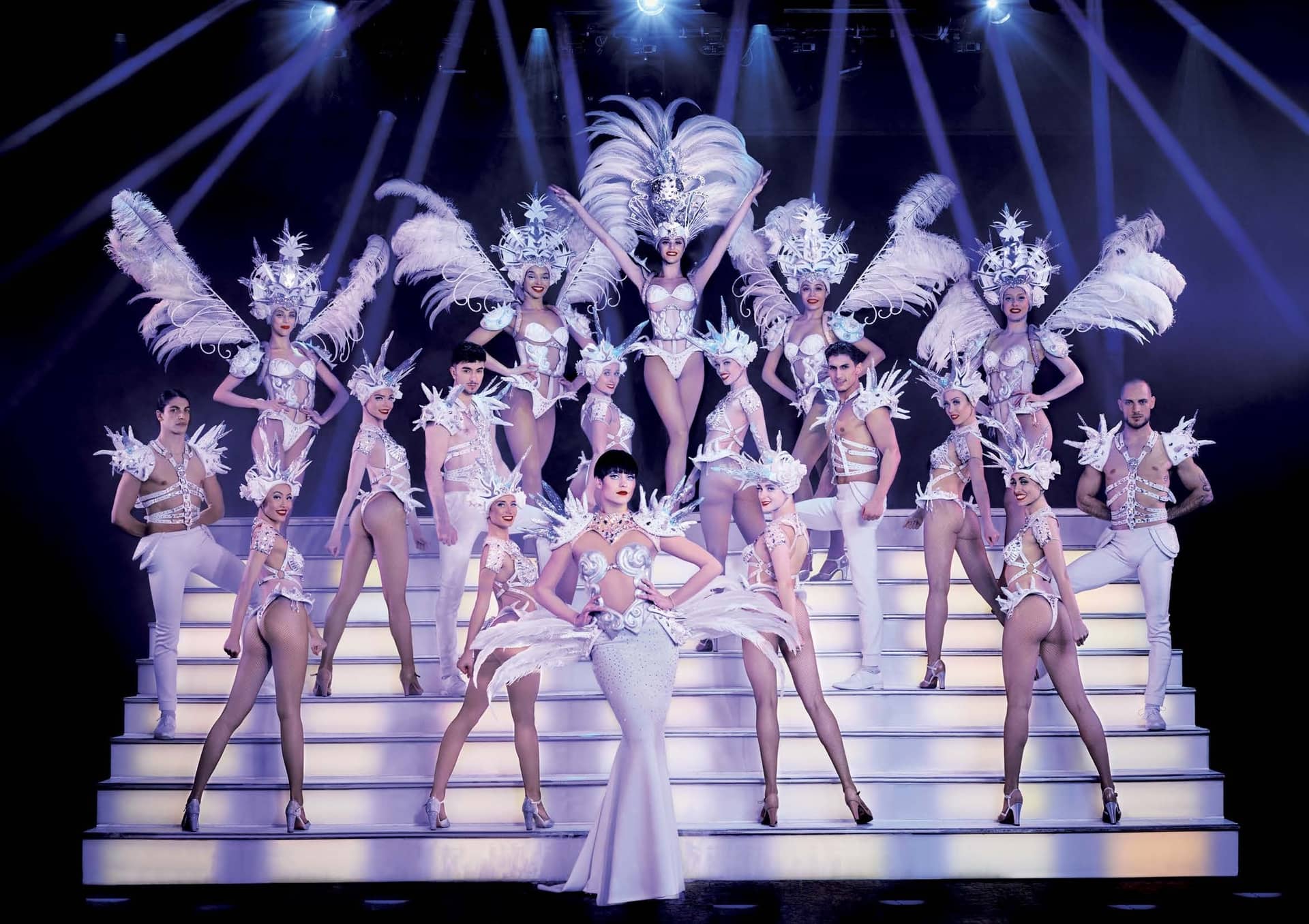 Troupe de revue en costumes blancs à plumes posant sur un grand escalier lumineux