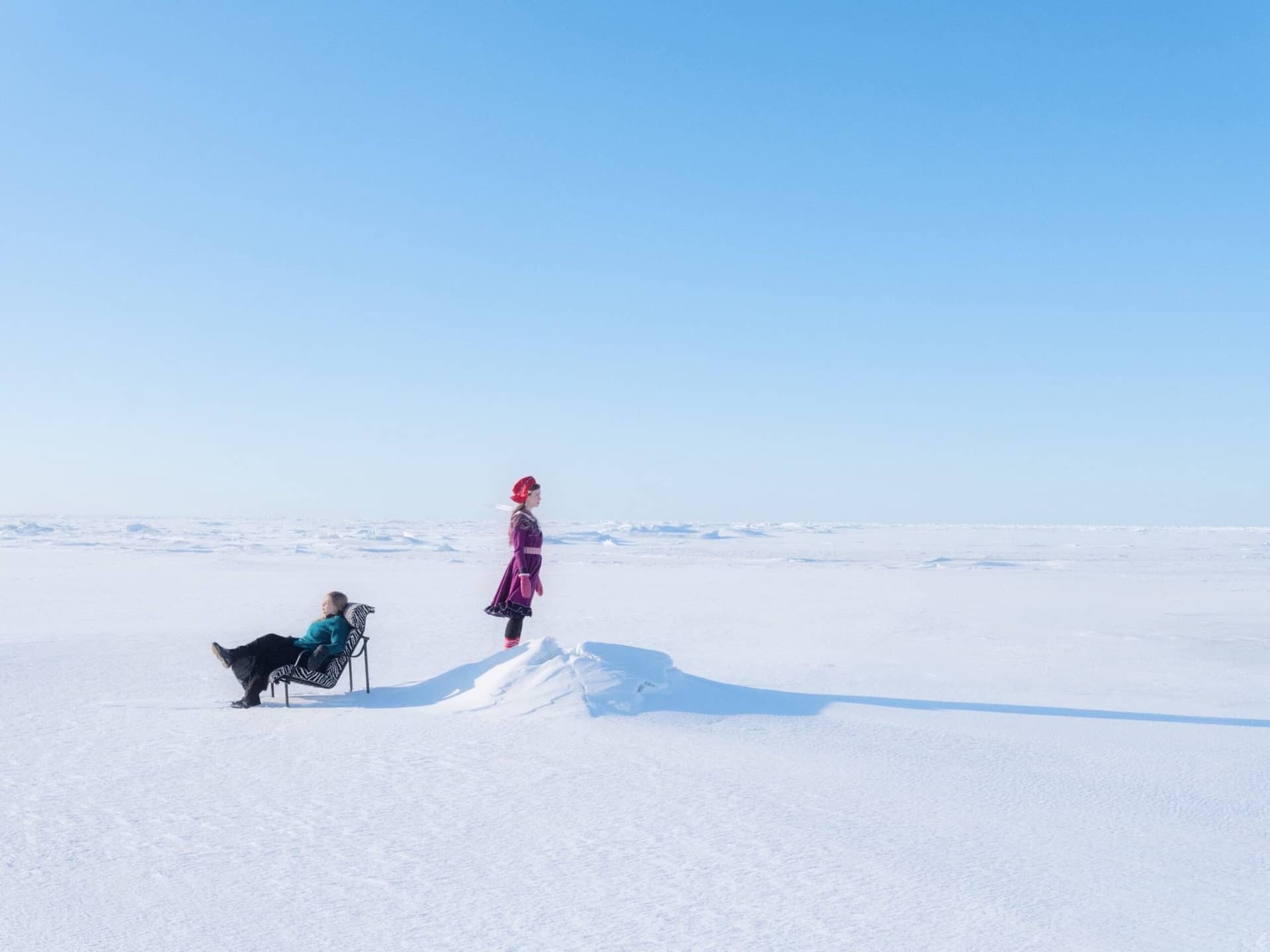 Deux figures en costume sámi traditionnel, l'une allongée sur un transat, au milieu d'une étendue de neige sous un ciel bleu