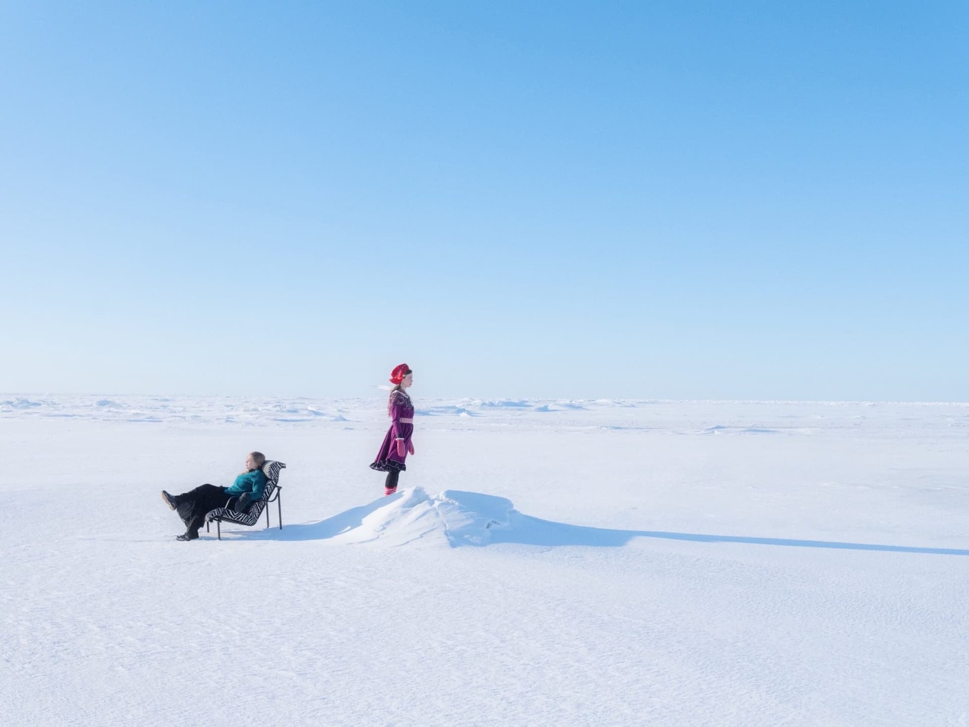 Deux figures en costume sámi traditionnel, l'une allongée sur un transat, au milieu d'une étendue de neige sous un ciel bleu