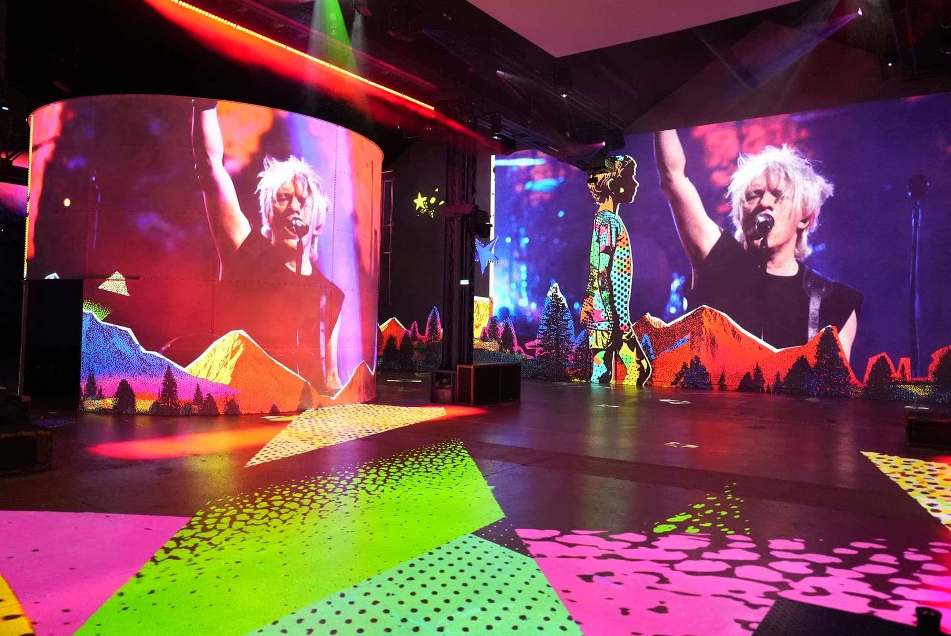 Le chanteur d'Indochine, bras levé, projeté sur un mur courbe de l'Atelier des Lumières, sol couvert de motifs fluo