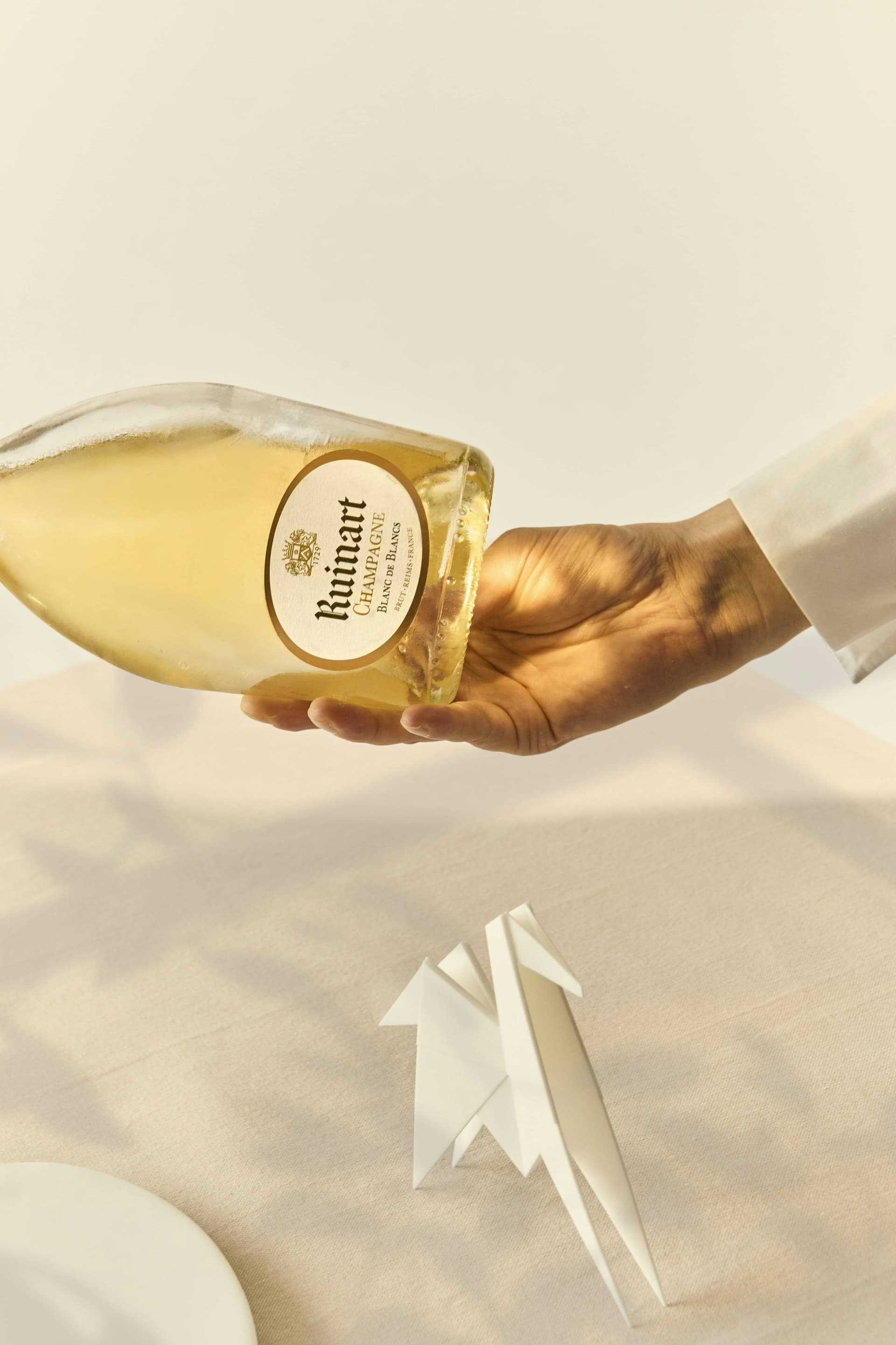 Main qui verse un champagne Ruinart Blanc de Blancs sur une nappe blanche, oiseau en papier plié