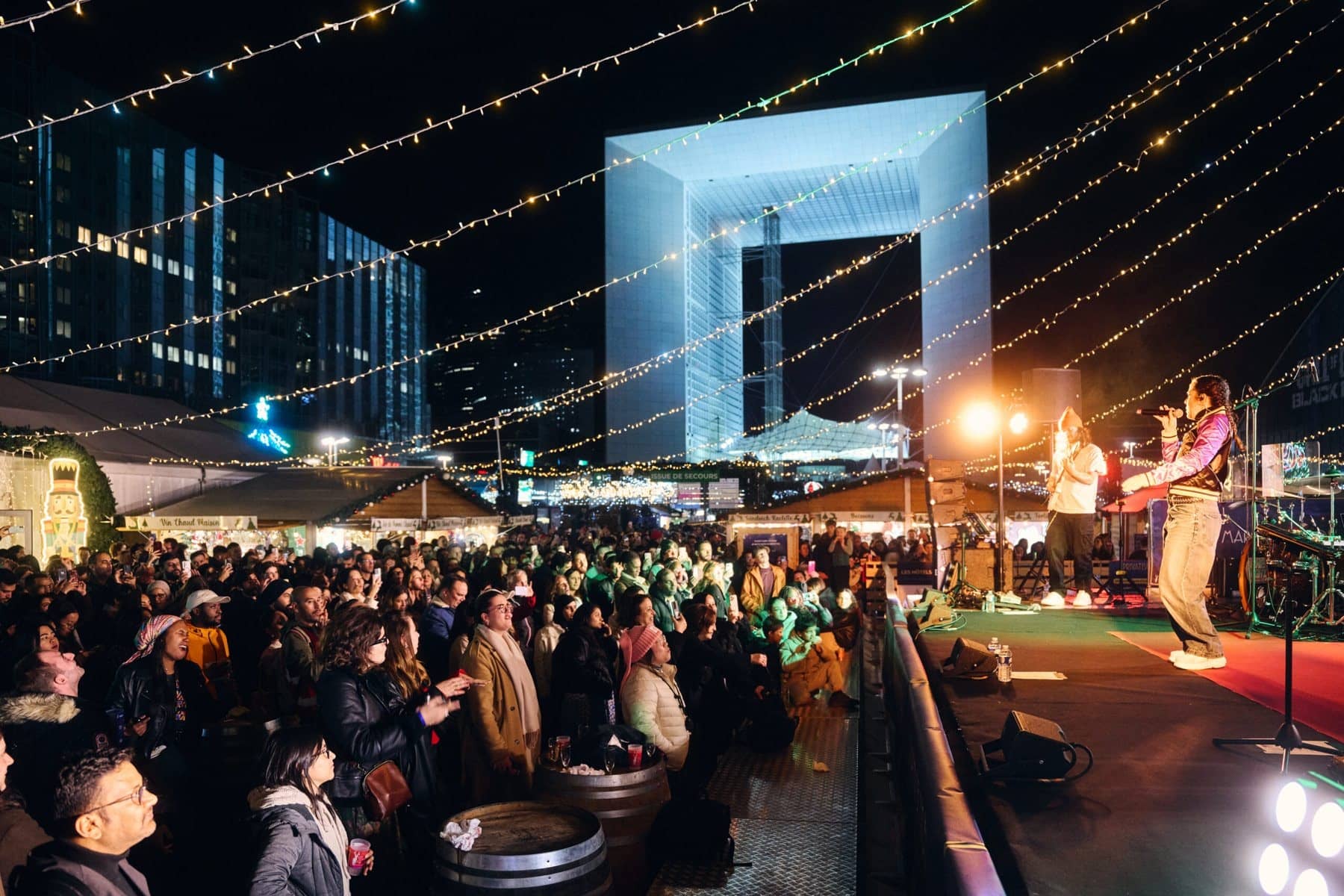 Crowd in front of a stage at the La Défense Christmas market, with the Grande Arche lit up behind