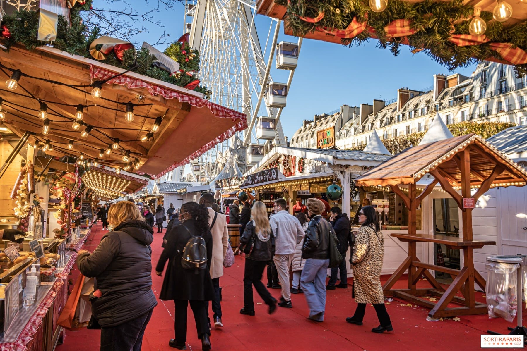 Lit wooden chalets and visitors on the red carpet of the Tuileries Christmas market, with the Ferris wheel behind