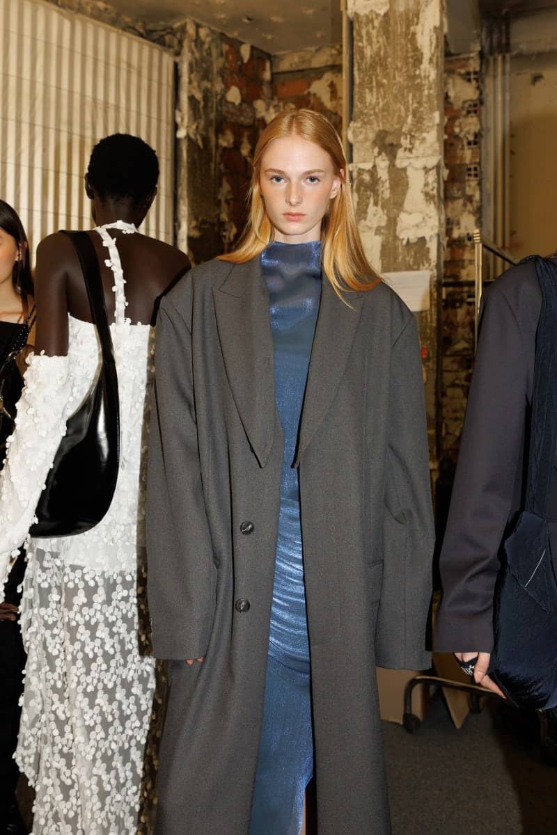 Mannequin en long manteau gris et robe bleue satinée en coulisses d'un défilé Re Rhée