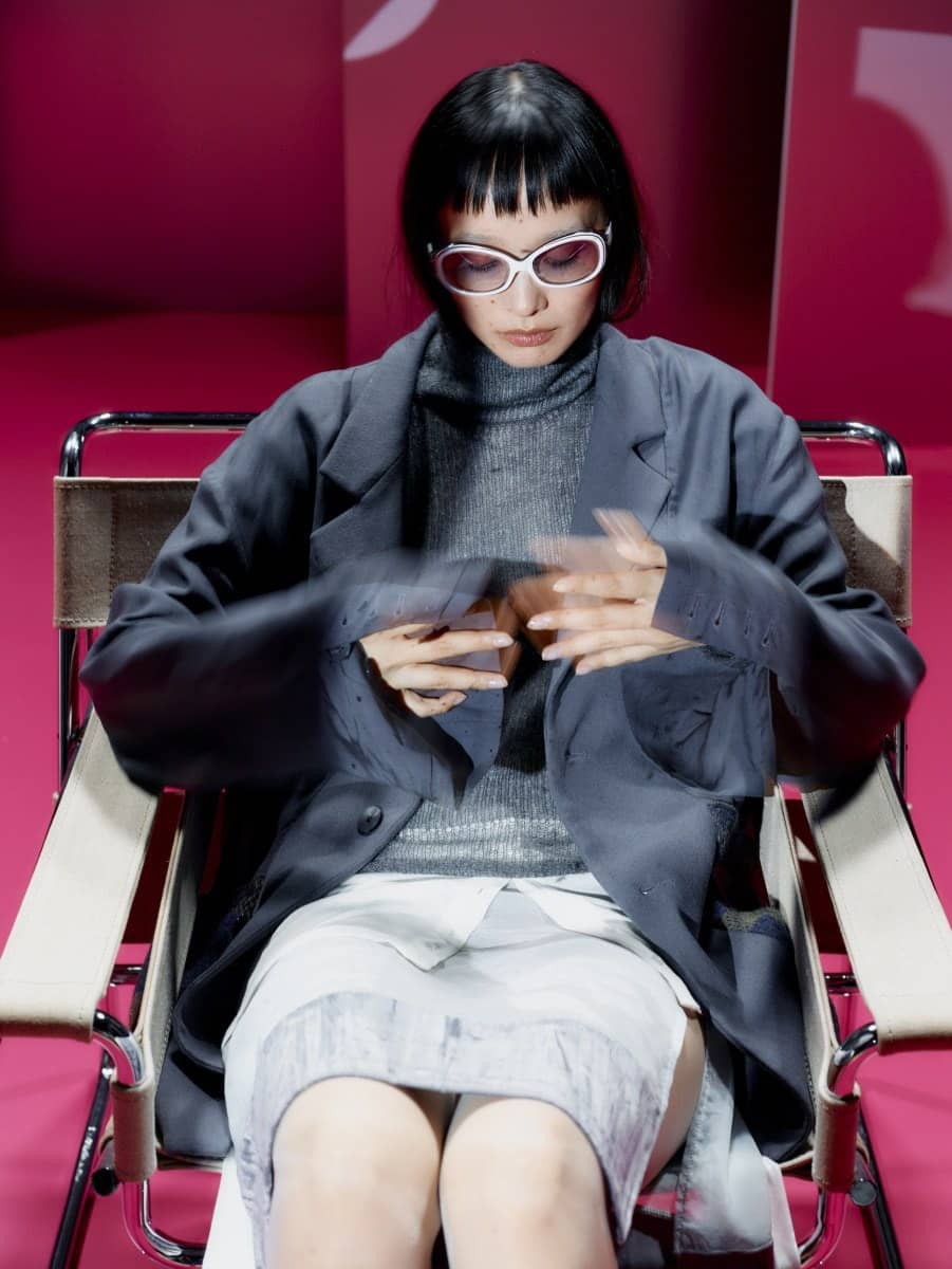 Mannequin en tailleur gris et col roulé assise sur une chaise en métal devant un mur rose, look Theilma