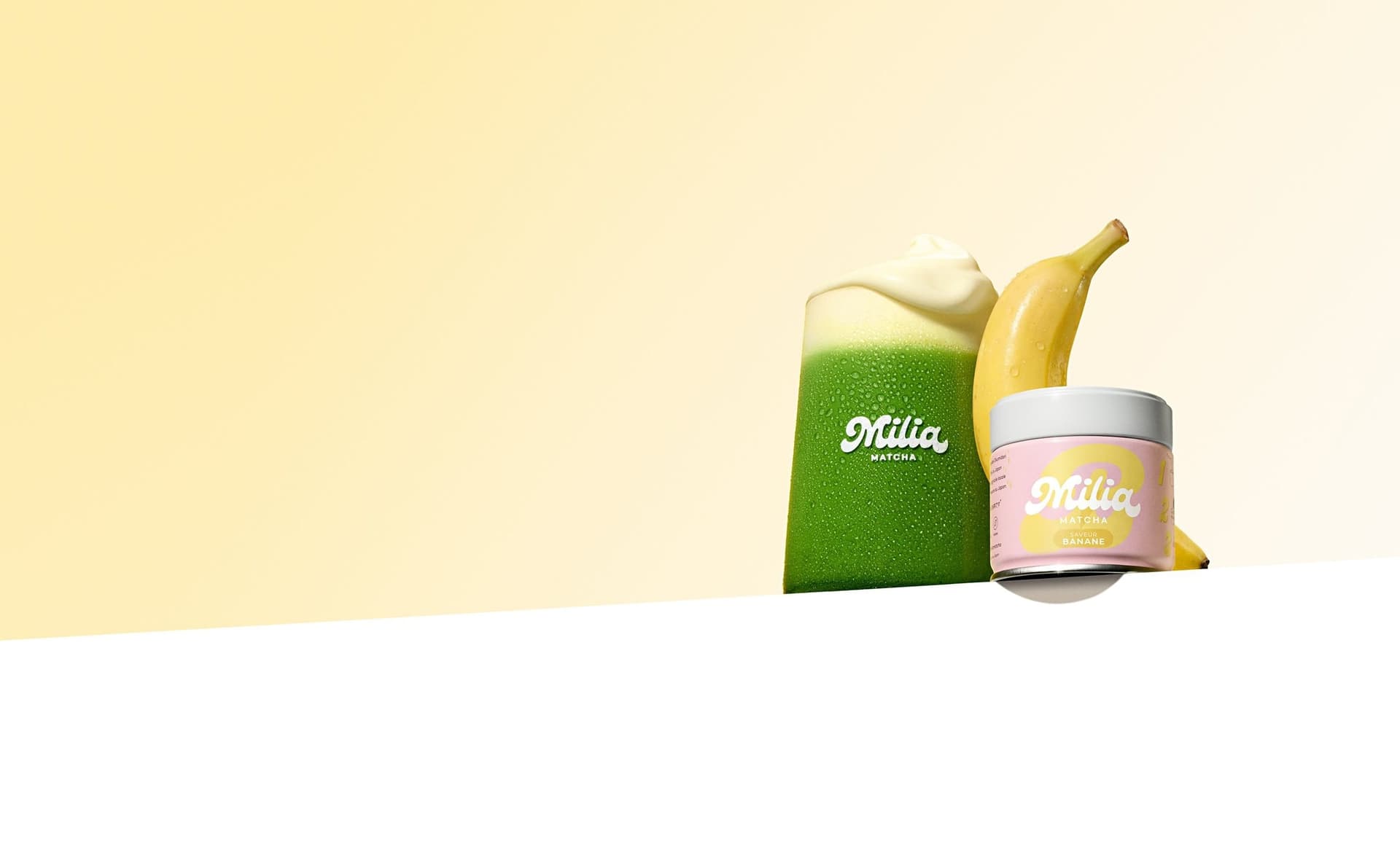 A glass of banana matcha topped with cream, a banana and a pink Milia Matcha tin on a pale yellow background
