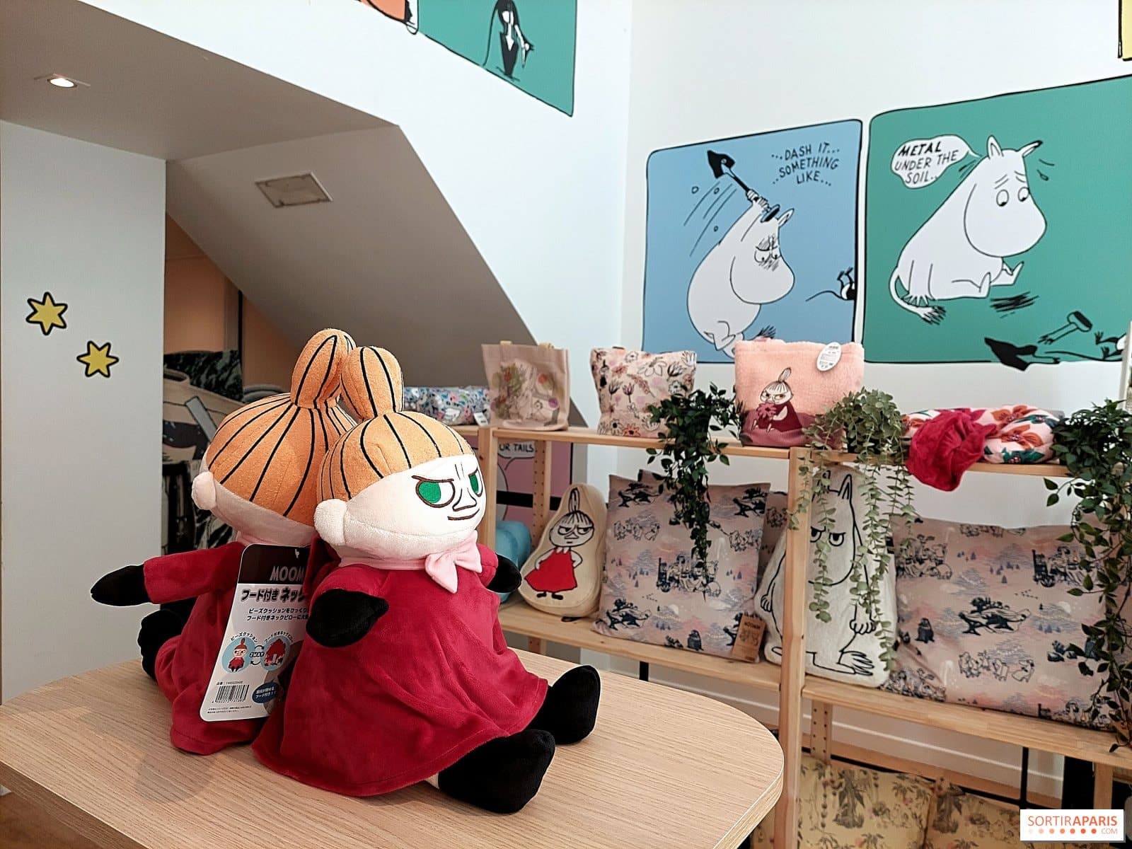 Peluche Petite My en robe rouge posée sur une table devant des affiches Moomin illustrées