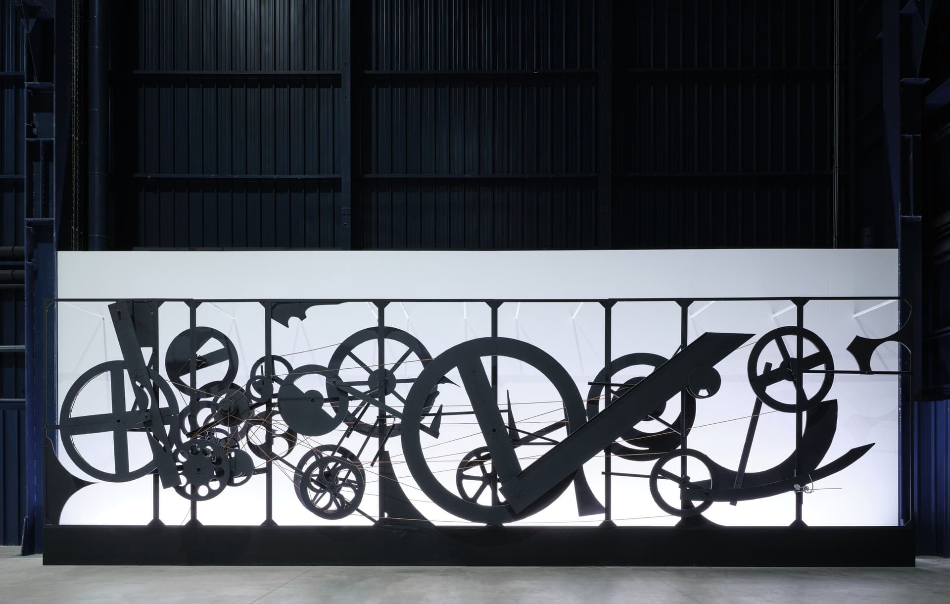 Requiem pour une feuille morte de Jean Tinguely, roues et engrenages noirs sur un long panneau lumineux blanc