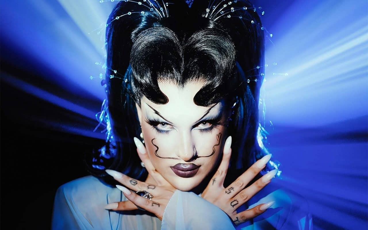 Portrait de la drag queen HitsuBlu, coiffure noire sculptée et maquillage graphique, sur fond de faisceaux bleus