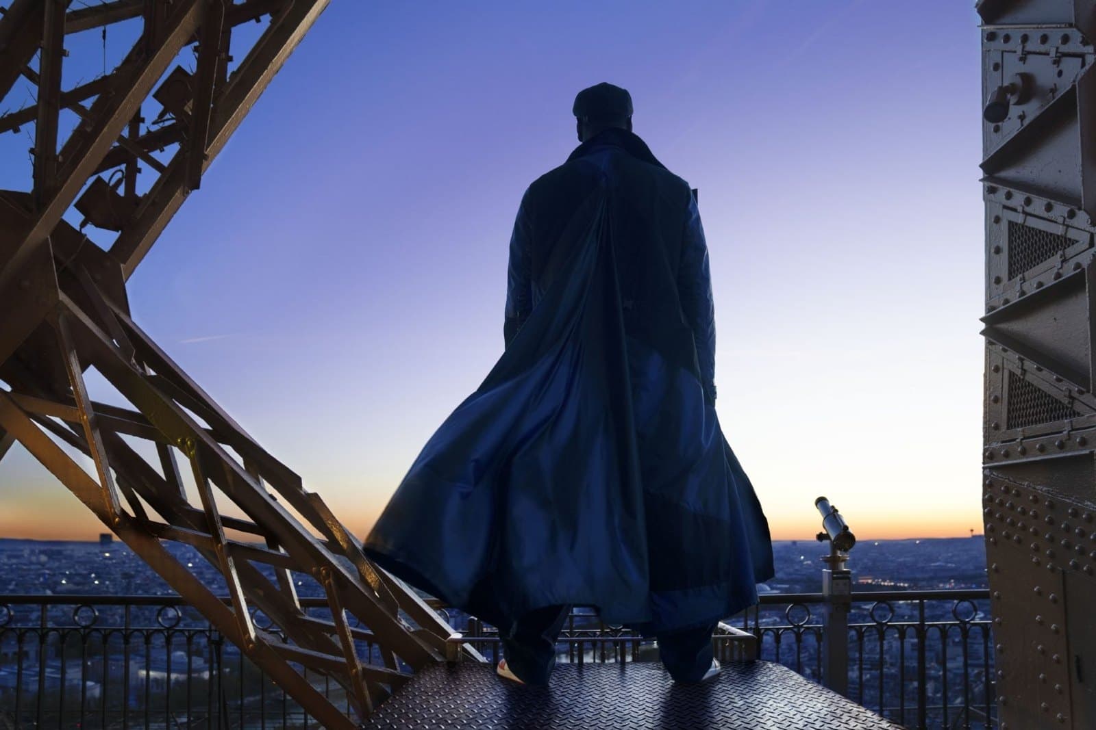 Assane Diop de dos, cape au vent, sur une plateforme de la tour Eiffel au crépuscule, dans la série Lupin