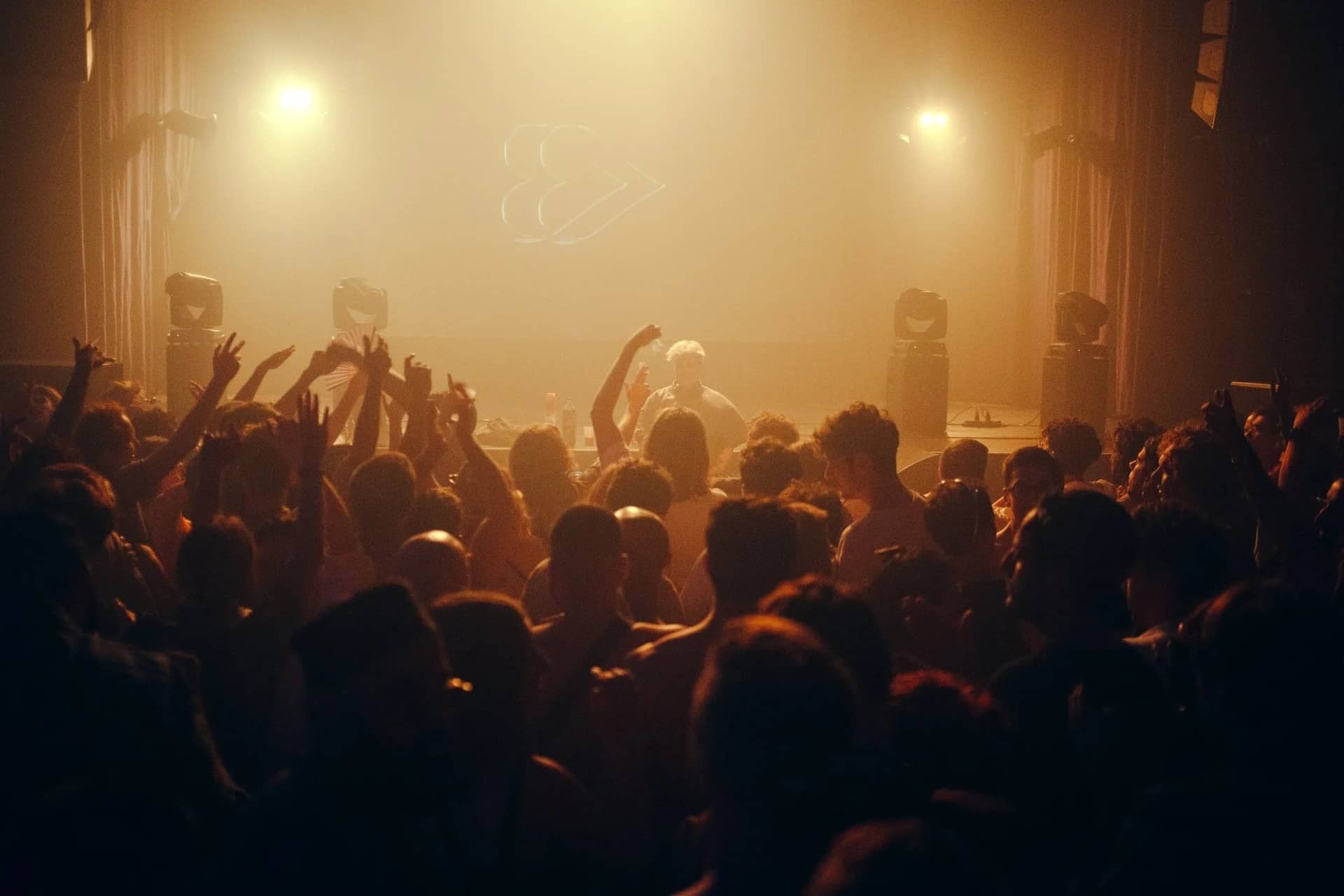 Foule bras levés face aux platines dans une salle enfumée baignée de lumière orange
