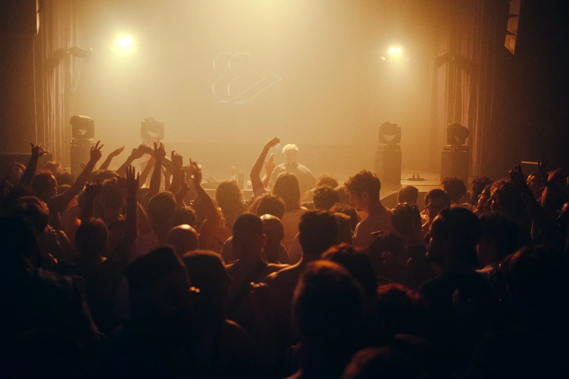 Foule bras levés face aux platines dans une salle enfumée baignée de lumière orange