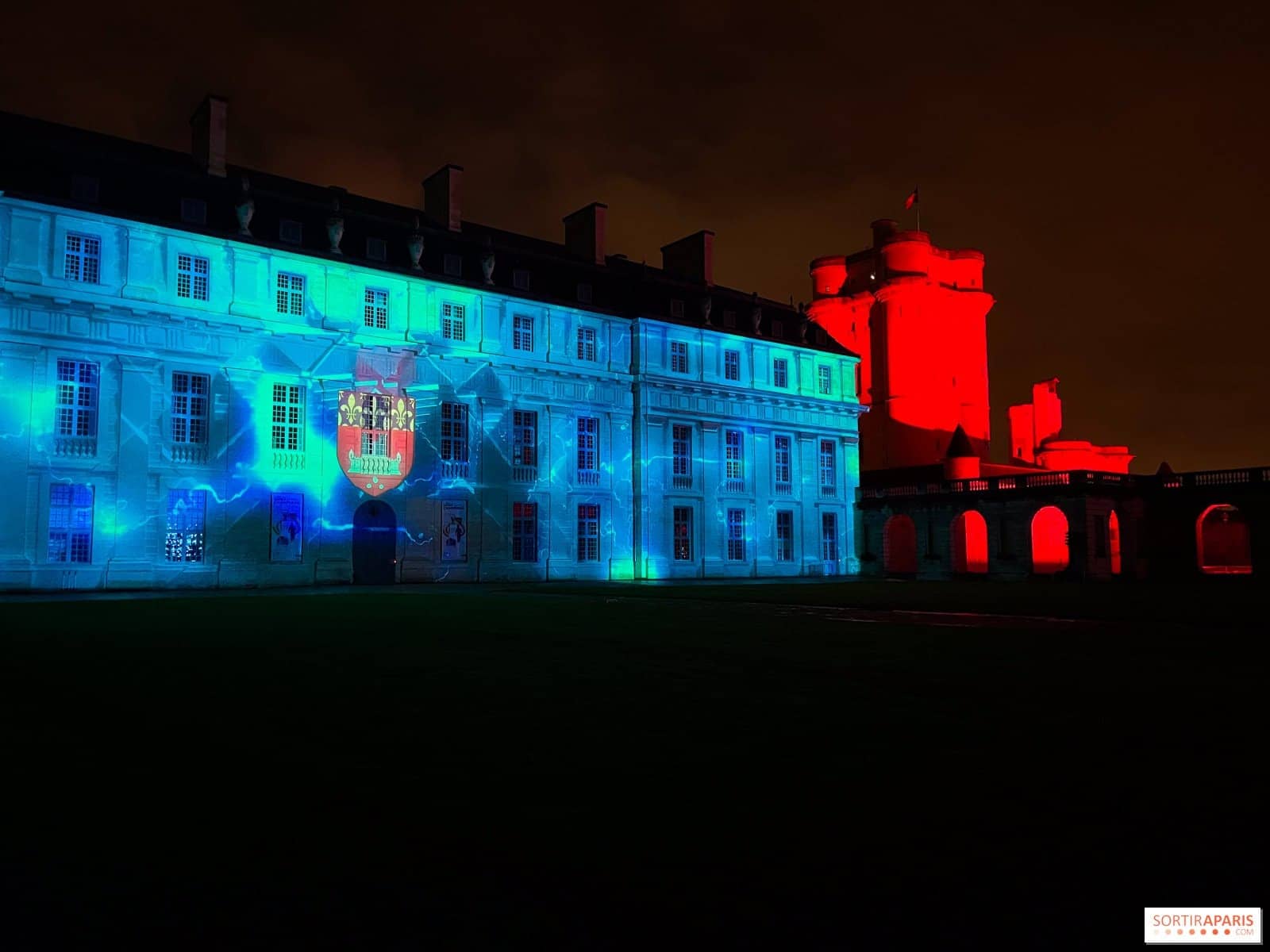 Pavillon du château de Vincennes éclairé en bleu, avec le donjon rouge en arrière-plan la nuit