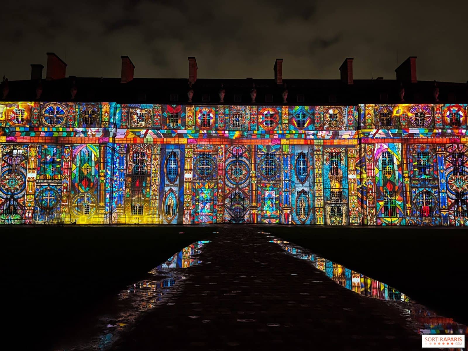 Façade du château de Vincennes couverte d'un mapping coloré, reflétée dans les flaques de la cour