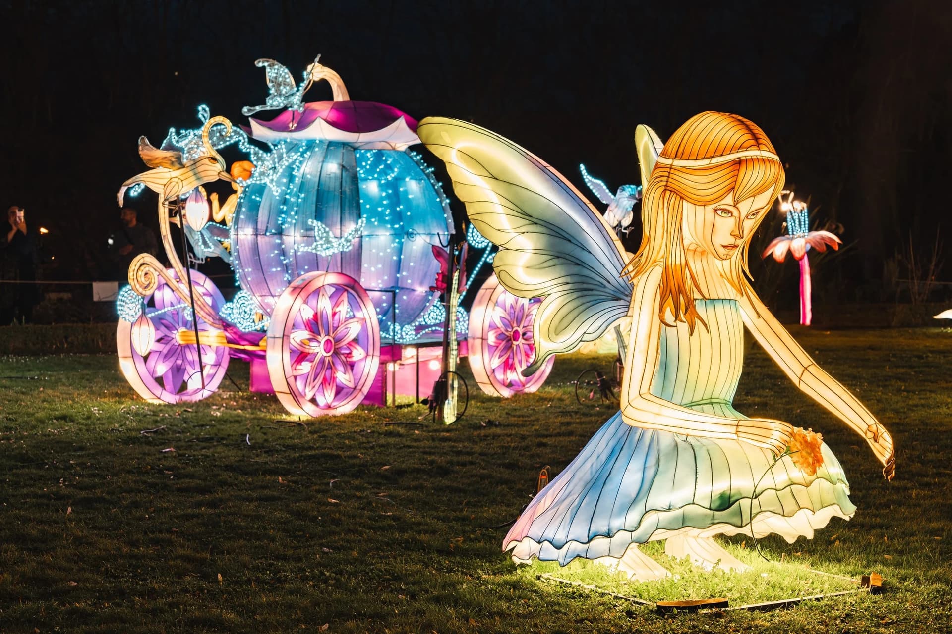 Lanterne géante d'une fée agenouillée devant un carrosse lumineux, sur la pelouse d'un parc la nuit
