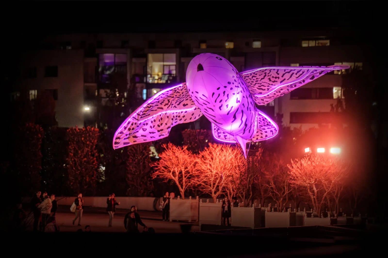 Hibou lumineux géant rose et violet posé au-dessus des haies du parc la nuit