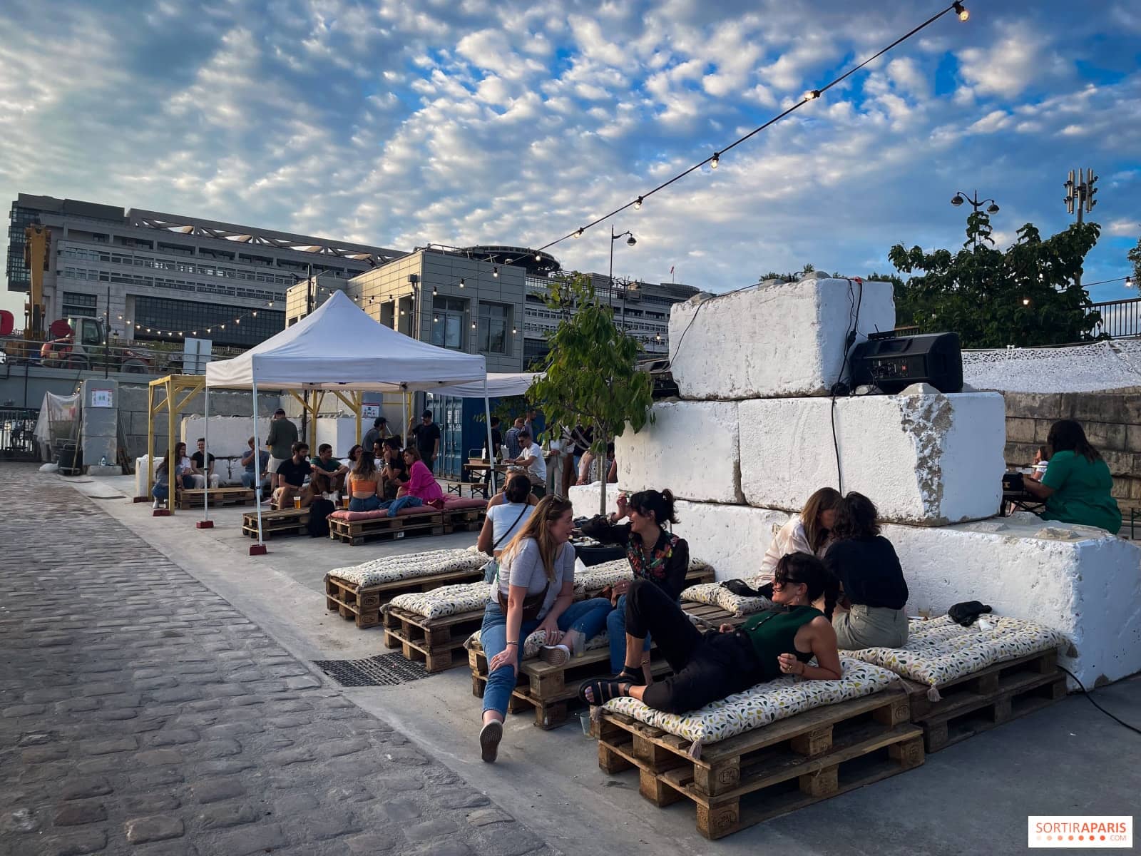 Blocs de ciment blancs réutilisés en banquettes avec coussins fleuris et palettes en bois, foule détendue au crépuscule à Quai Sauvage Bercy