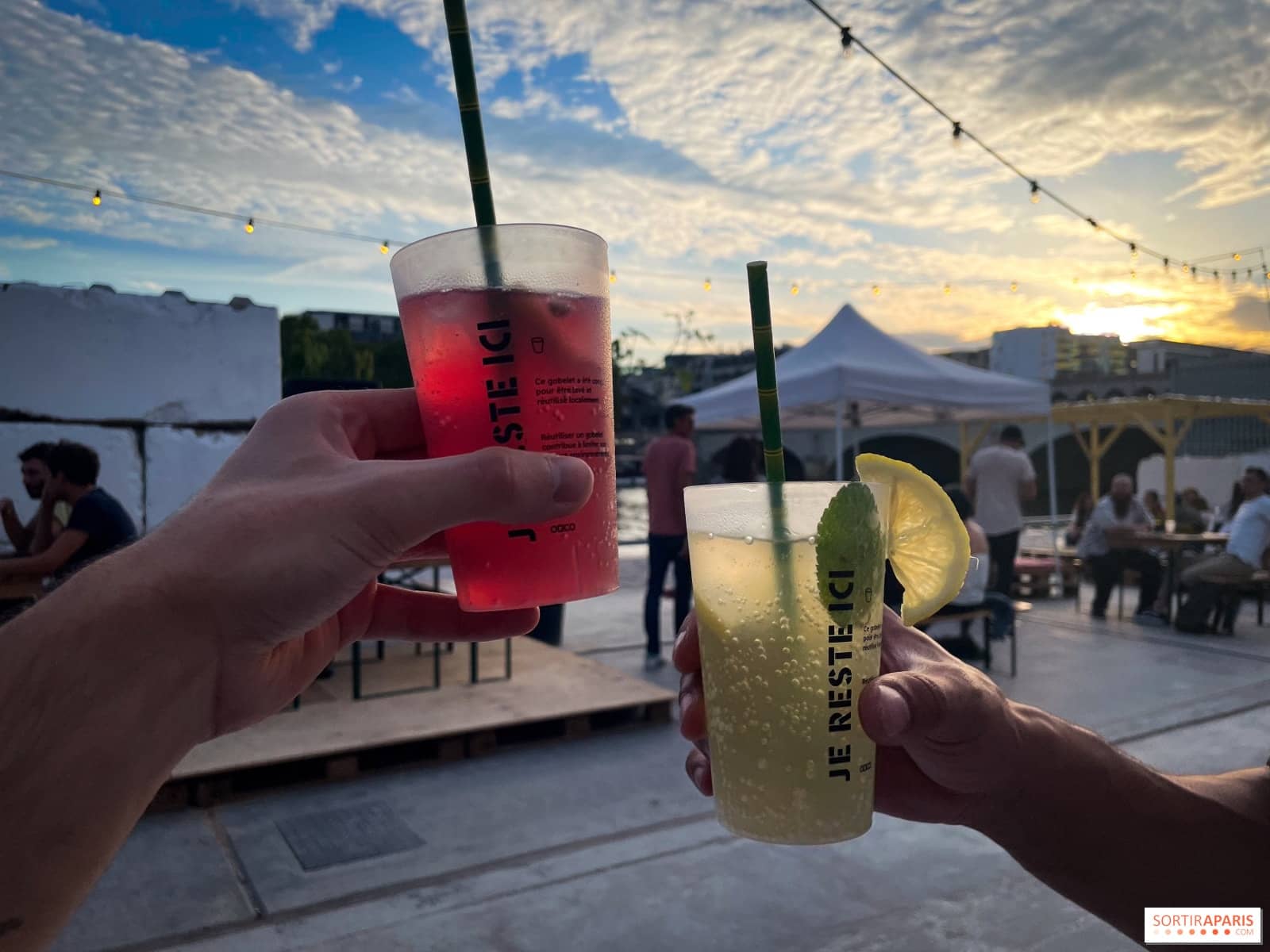 Deux cocktails dans des gobelets réutilisables marqués JE RESTE ICI tenus à bout de bras au-dessus du quai au sunset