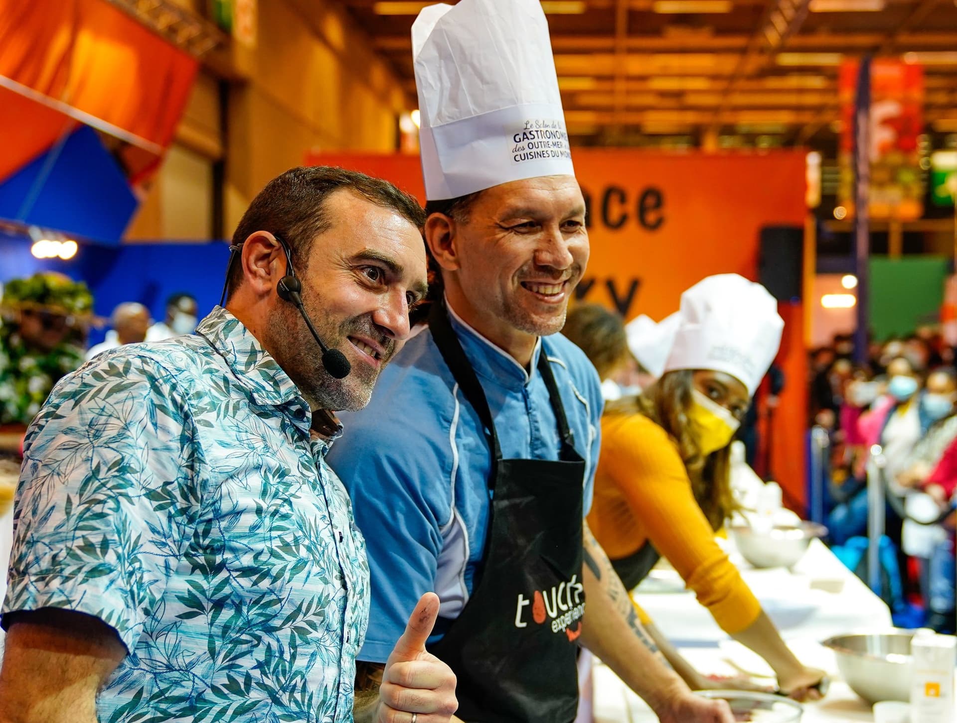 Deux chefs en démonstration culinaire au micro sur un stand coloré du salon Sagasdom