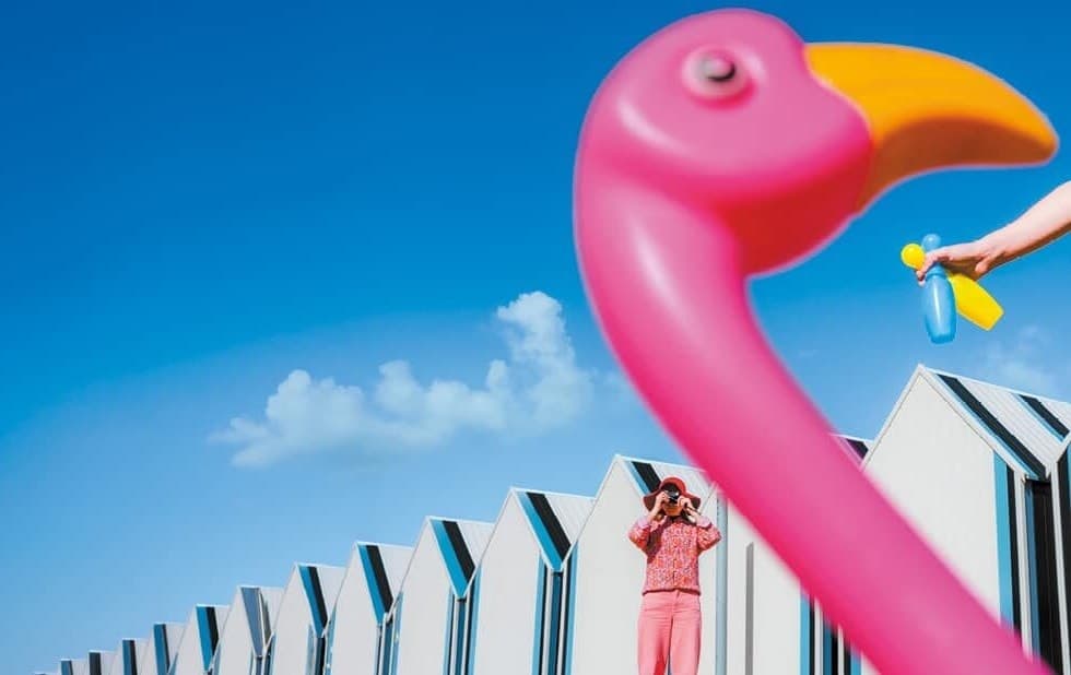 Flamant rose gonflable devant des cabines de plage blanches, une photographe en rose vise l'objectif
