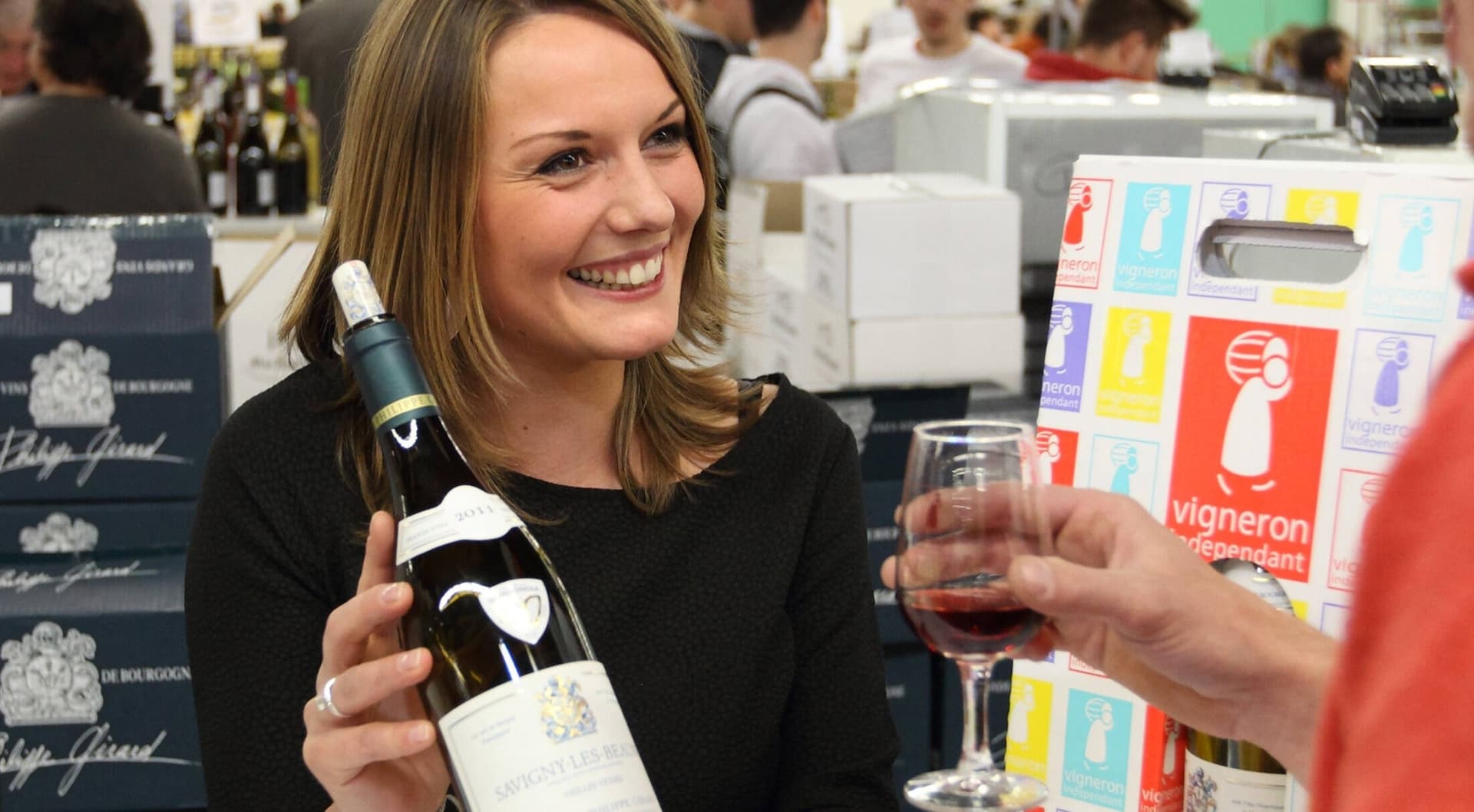Une vigneronne souriante présente une bouteille à un visiteur sur son stand d'un salon des Vignerons Indépendants