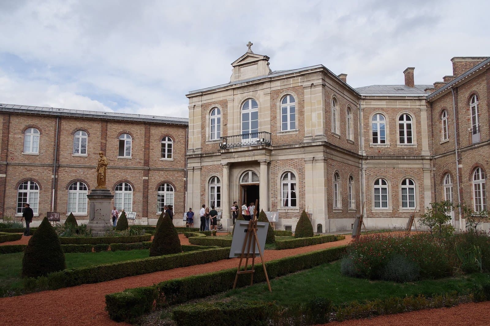 The inner garden of the Fondation Eugène Napoléon, clipped beds and a gilded statue in front of the brick and stone facade