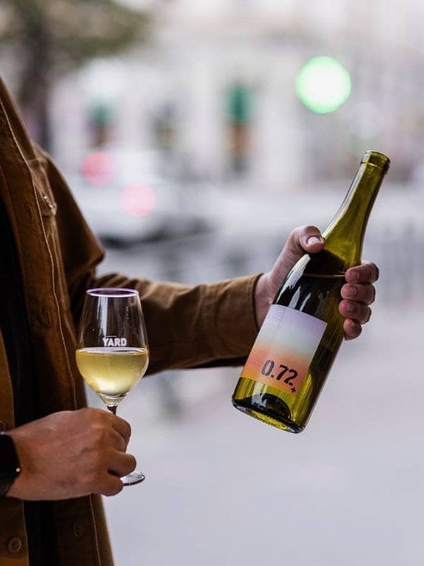 Une main tient un verre gravé YARD rempli de blanc et une bouteille de vin nature dans la rue